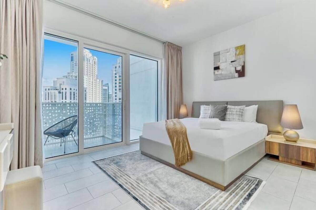 Un dormitorio moderno con una cama grande, vistas a la ciudad a través de ventanales y un sillón, además de una mesita de noche con lámparas.