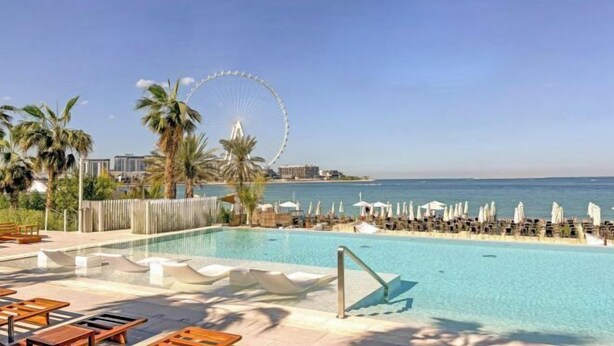 Ein Poolbereich mit Liegestühlen und Sonnenschirmen, im Hintergrund ein Riesenrad und freie Sicht auf das Meer.