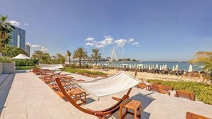 Eine Strandterrasse mit Holzliegen und Sonnensegel, mit Blick auf das Meer und ein ferner gelegenes Riesenrad.