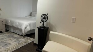 Un dormitorio con cama, mesita de noche, lámpara, pequeña mesa con un objeto decorativo, silla y alfombra.