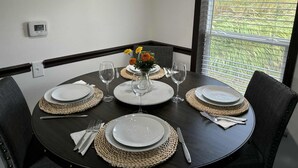 Una mesa de comedor dispuesta con platos, vasos y cubiertos, rodeada de sillas, con un termostato y una ventana con persianas en el fondo.