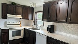Una cocina con gabinetes de madera oscura, una encimera blanca, una campana extractora de acero inoxidable, un horno empotrado, un lavavajillas y una cafetera.