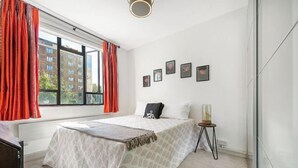 Une chambre avec un grand lit, une fenêtre avec des rideaux rouges, une table de chevet avec une lampe et des tableaux encadrés accrochés au mur.