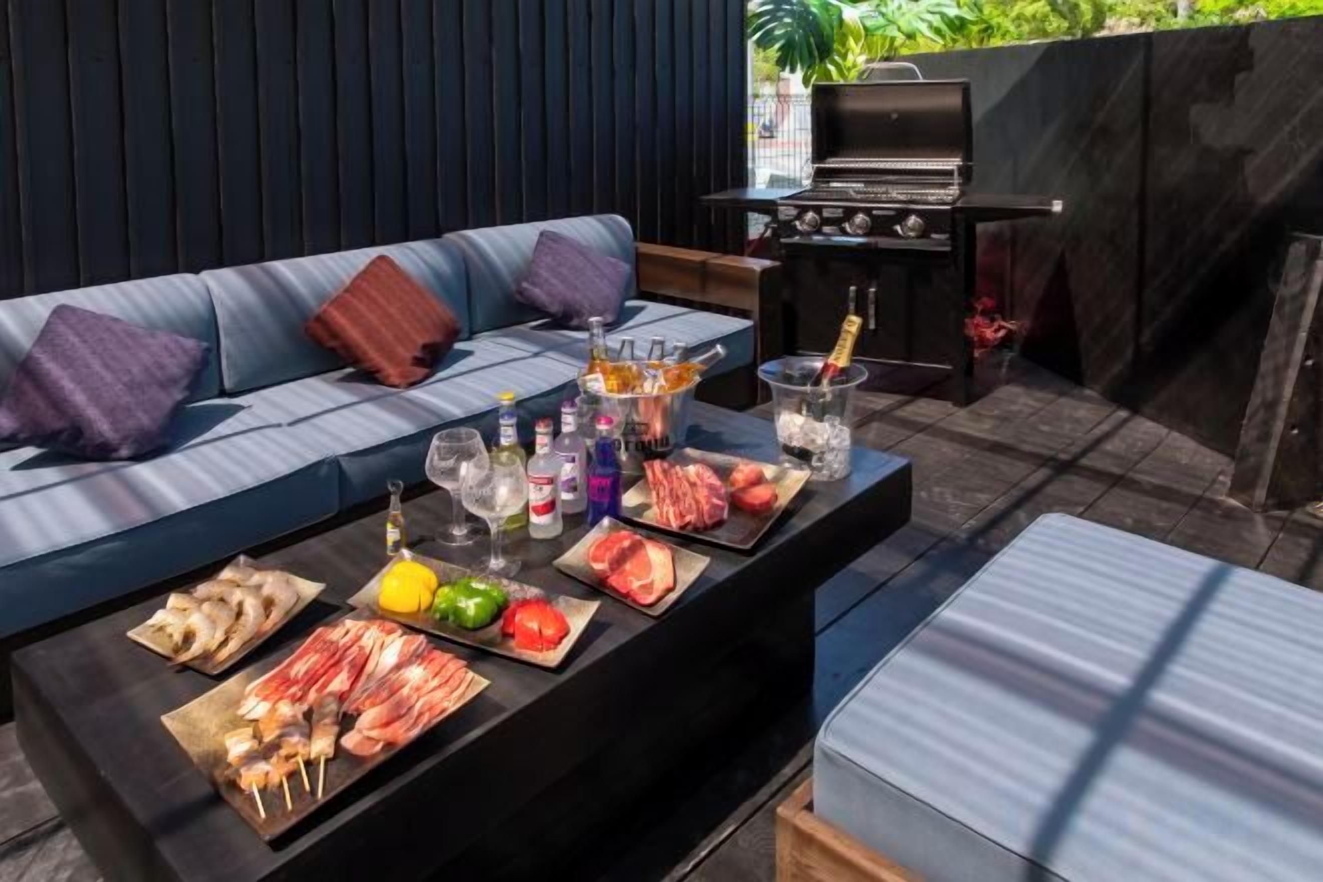 An outdoor seating area with a barbecue grill, a sofa, and a table with food and drinks.