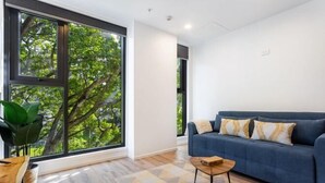 A modern living room with a blue sofa, a small wooden coffee table, and a large window with a view of trees. - Sunny Retreat in Heart of CBD (Auckland)