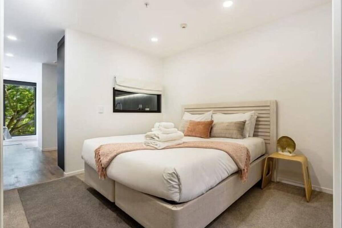 A modern bedroom with a large bed, a bedside table, a small stool, and a wall-mounted TV.