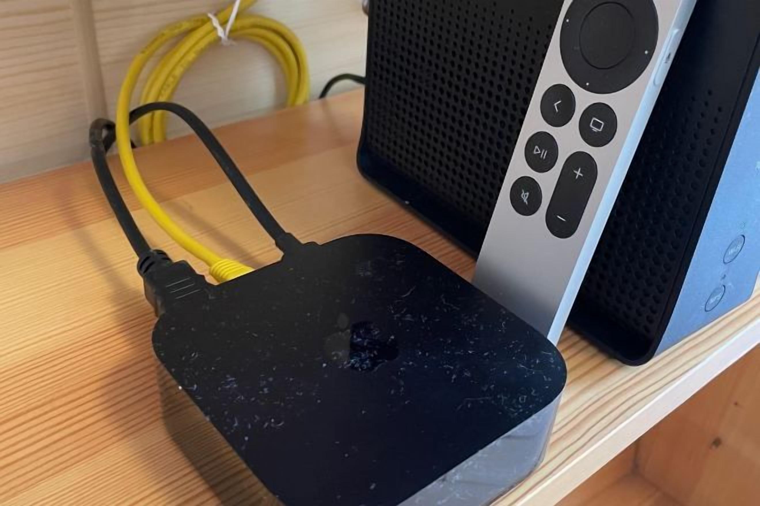 A black, damaged router with yellow cables on a wooden surface, next to a silver speaker with a remote control.