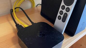 A black, damaged router with yellow cables on a wooden surface, next to a silver speaker with a remote control.
