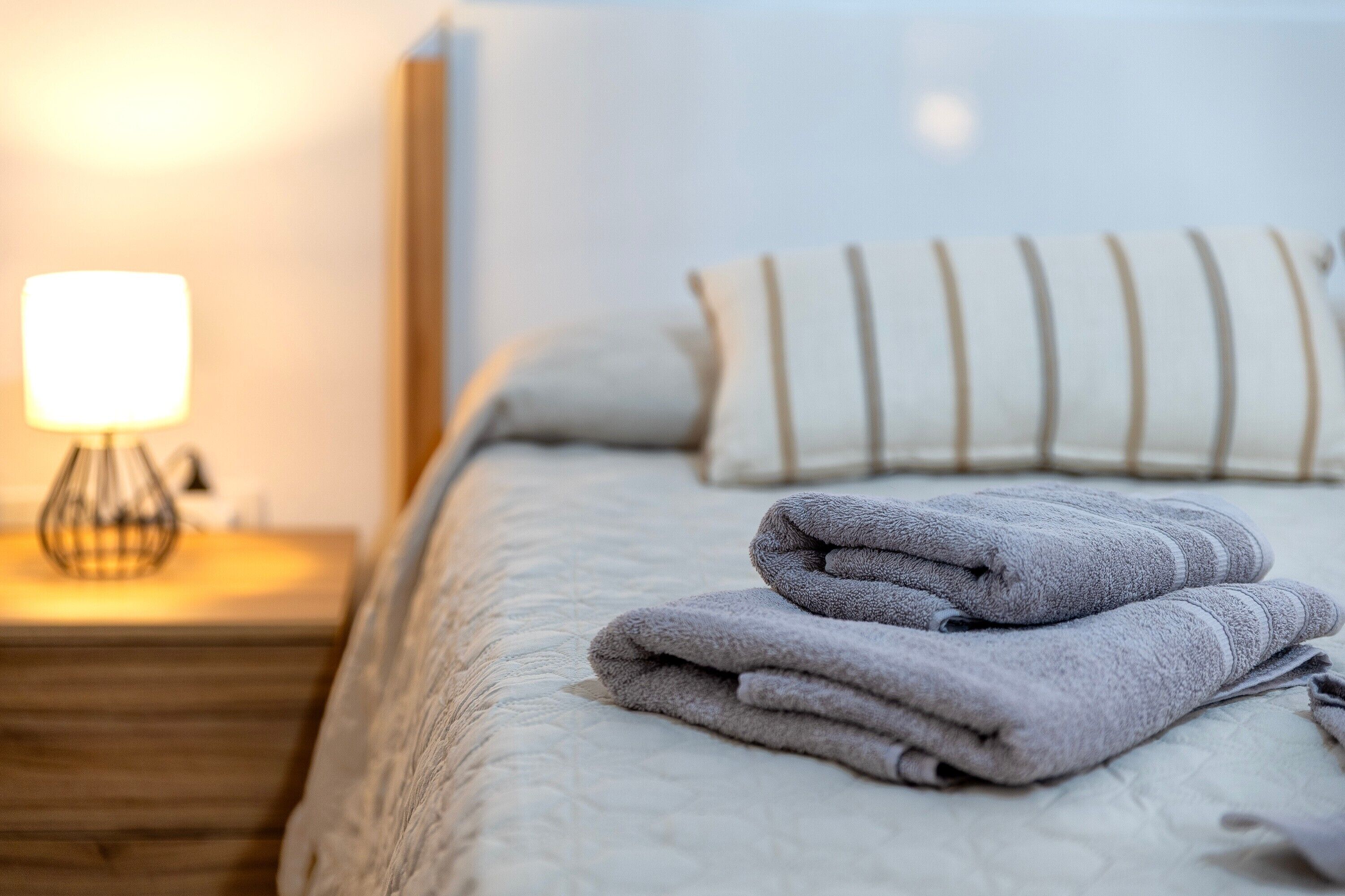Una cama bien hecha con toallas dobladas, una lámpara de noche y un cojín a rayas.