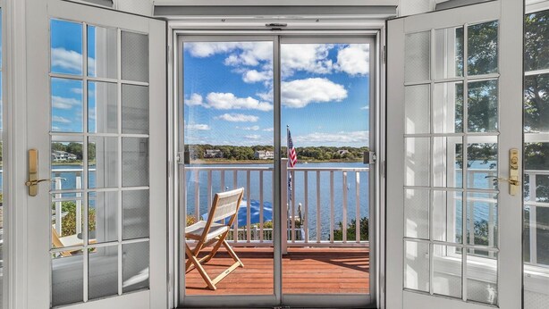 Vue depuis une pièce dont la porte est en verre et qui donne sur une terrasse avec une chaise, d’où l’on aperçoit un lac et des maisons.