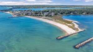 Vue aérienne d’une zone côtière avec une plage de sable, des eaux d’un bleu limpide et une zone résidentielle avec des maisons et de la végétation en arrière-plan.
