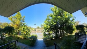 A covered outdoor area with a pathway leading to a water feature, surrounded by lush greenery and a clear blue sky. - GuddahHomestay (Sawang Daen Din)