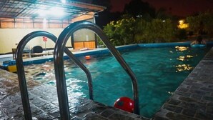 Una piscina con escaleras de metal y una pelota roja, rodeada de baldosas de piedra y un área cubierta con techo.