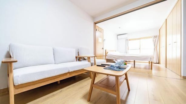 Habitación minimalista con suelo de madera, un sofá blanco, una mesita de madera y una cama con ventana.