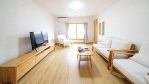 Una sala de estar con una televisión sobre un mueble de madera, un sofá blanco, una mesa de centro de madera y una silla.