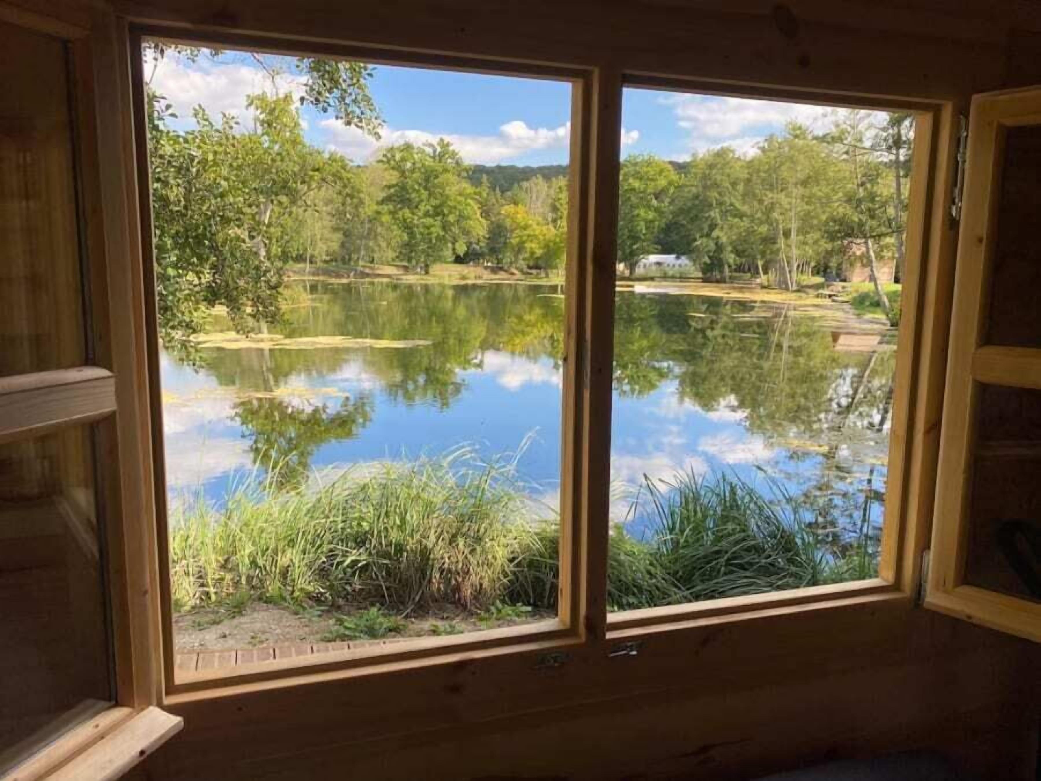 A wooden cabin with a large window overlooking a calm lake surrounded by trees.