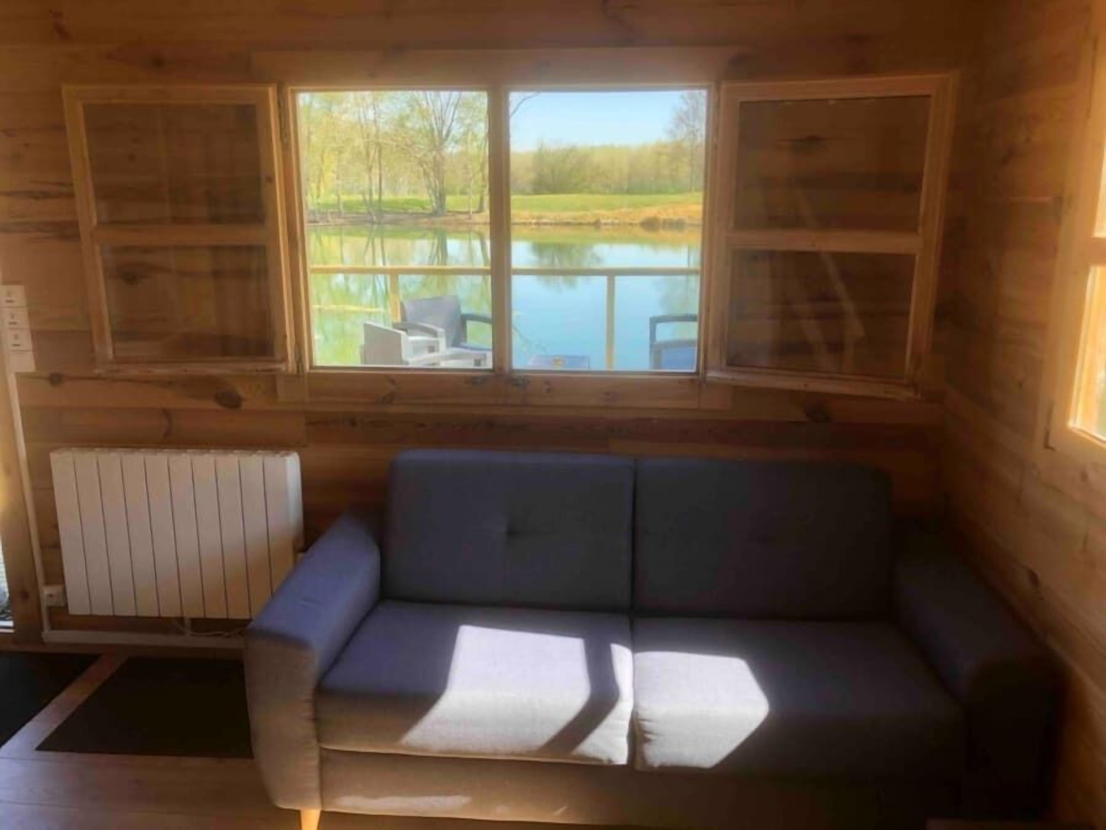 Interior de una cabaña de madera con un sofá, una ventana con vista a un lago y un radiador.