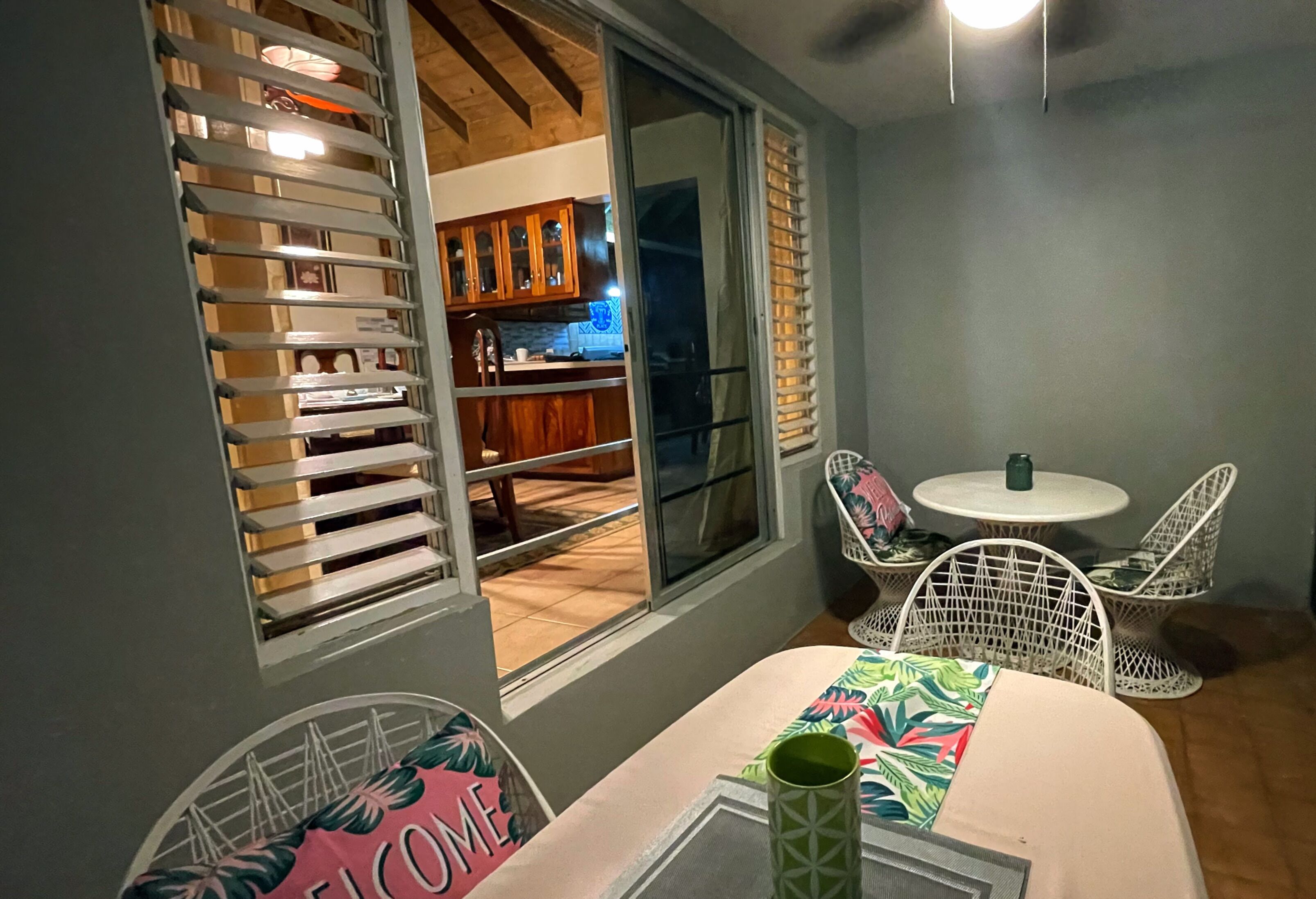 Habitación con comedor que incluye una mesa con un cartel de 'Welcome', sillas de mimbre blancas y vista a una cocina por un puerta corrediza de vidrio.