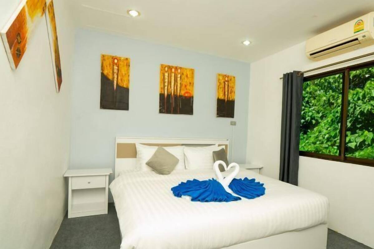 A neatly made bed with a blue towel and a white swan design, a white nightstand, a wall with three paintings, and a window with a view of greenery.