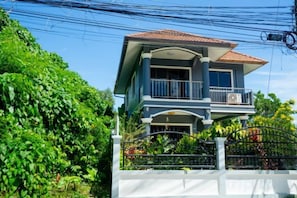 Una casa de dos pisos con balcón, rodeada de follaje y una cerca blanca.