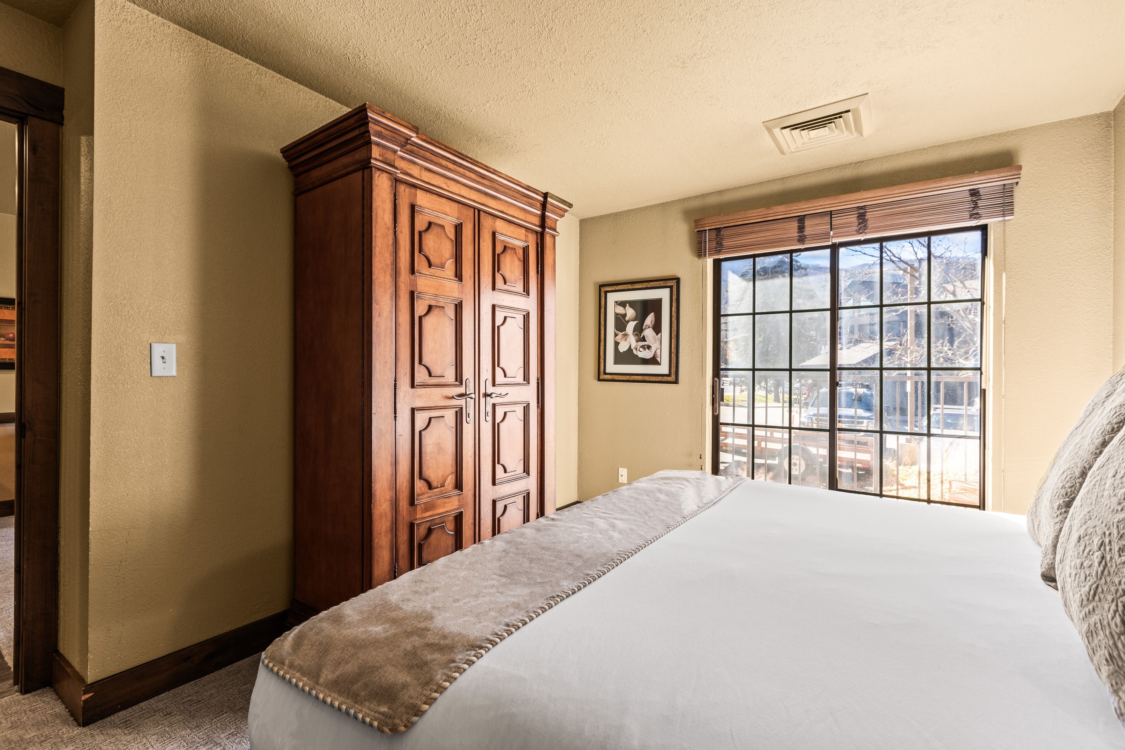A bedroom with a large bed, a wooden wardrobe, a window with blinds, and a framed picture on the wall.