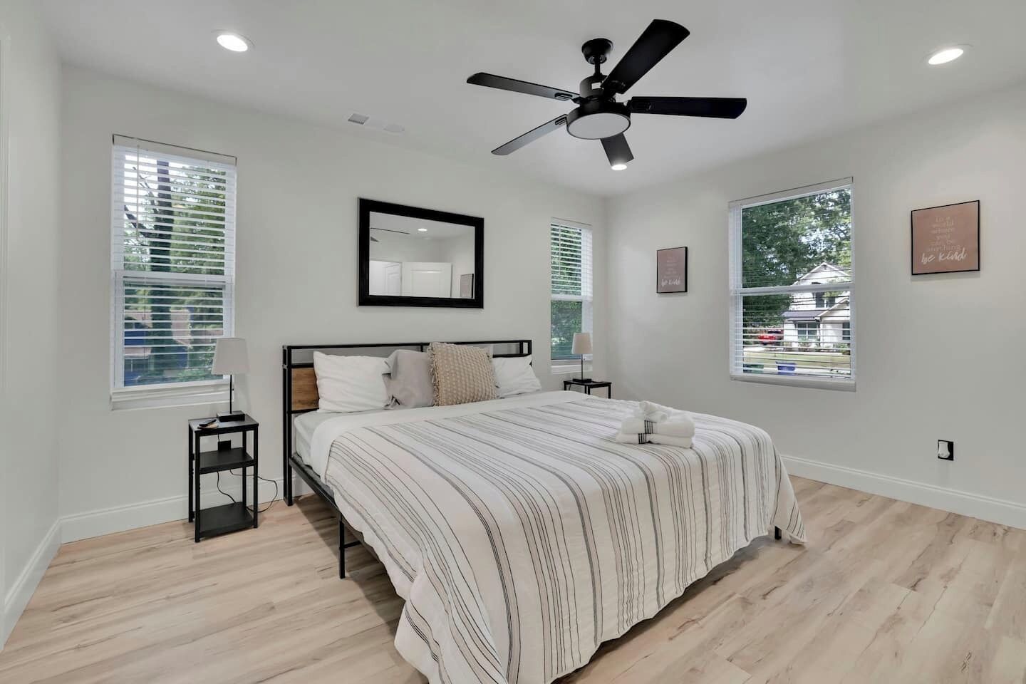 A bedroom with a bed, a ceiling fan, two bedside tables, a mirror, and two windows with blinds.