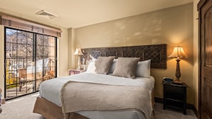 Ein Schlafzimmer mit einem großen Bett, einem Nachttisch mit Lampe, einem Fenster mit Jalousien und Blick ins Freie.