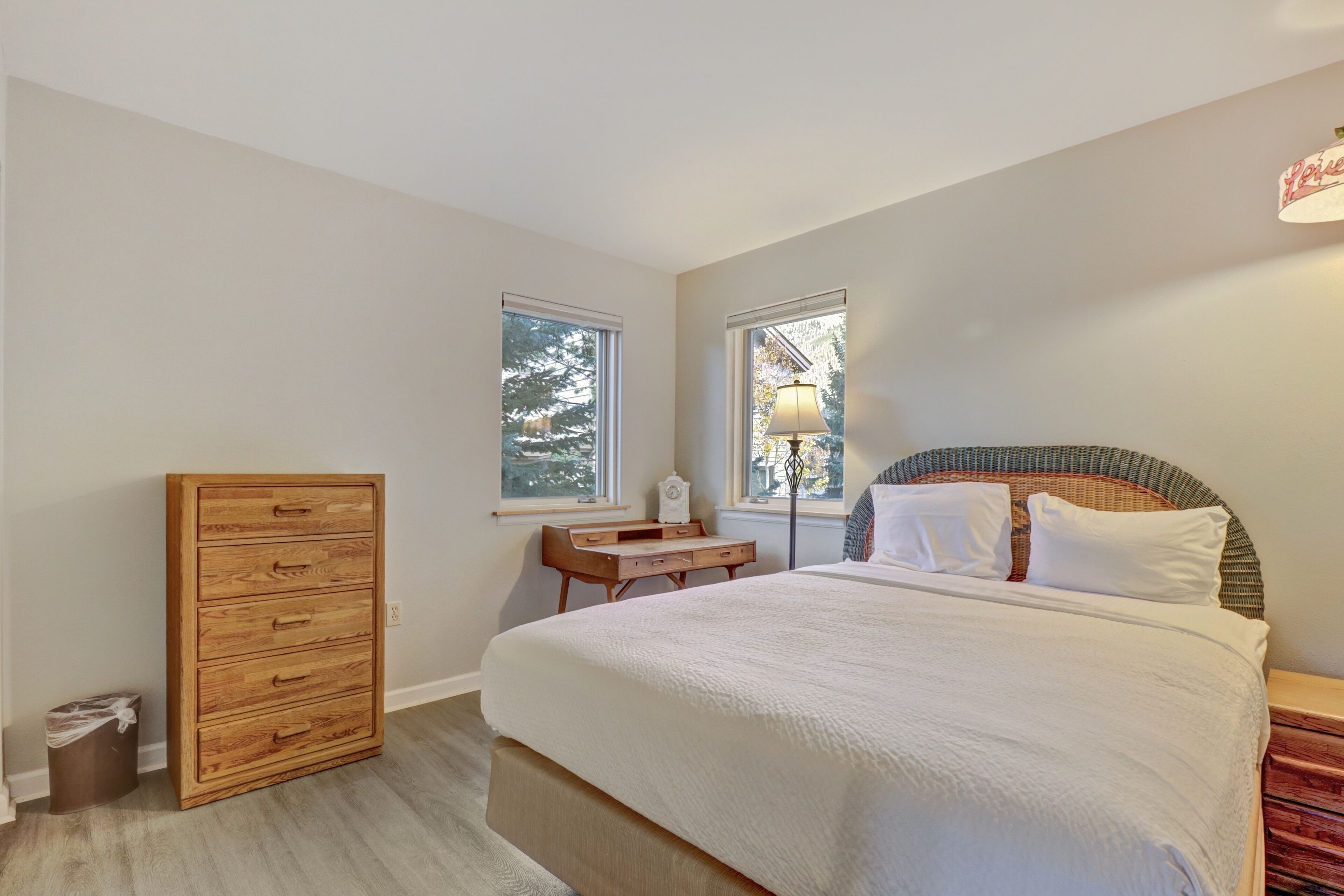 A bedroom with a bed, a wooden dresser, a desk, and two windows.