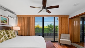 A hotel room with a large bed, a ceiling fan, a chair, and a view of greenery through the sliding glass door.