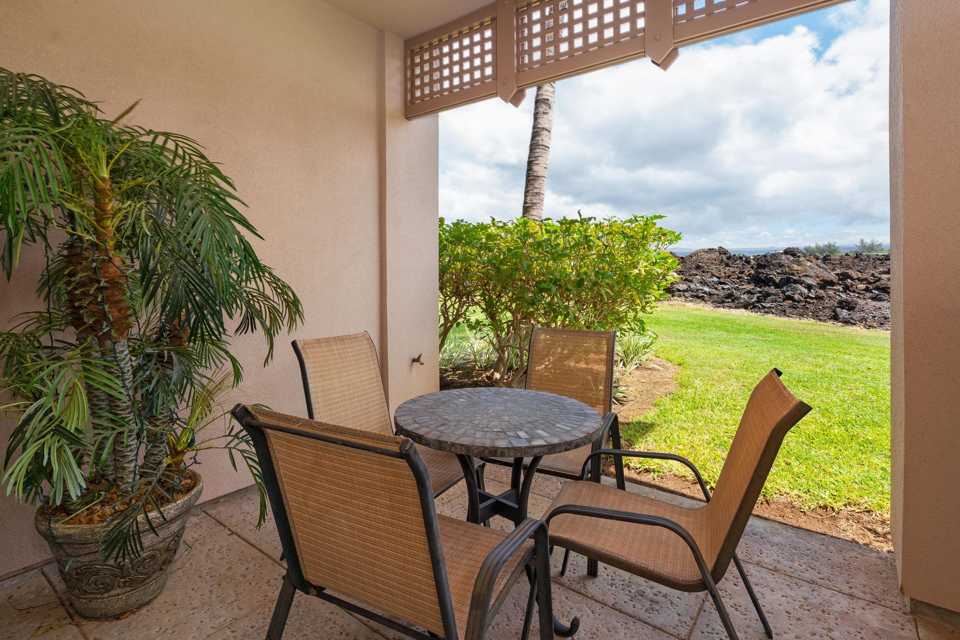 Un patio con una mesa redonda y cuatro sillas, una palmera en maceta y vistas a una zona verde con un paisaje rocoso al fondo.