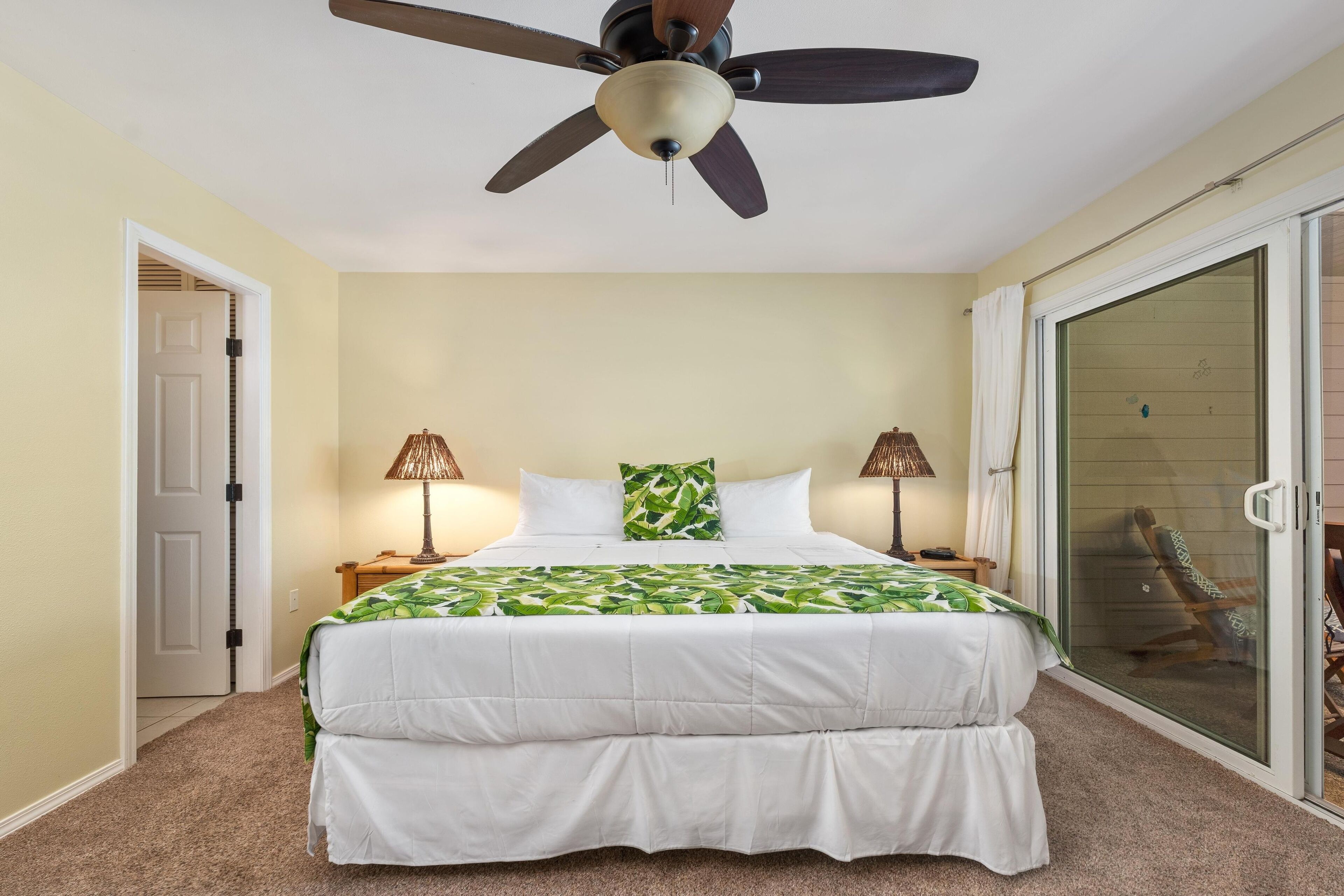 A bedroom with a bed, two bedside lamps, a ceiling fan, a sliding glass door, and a chair outside.