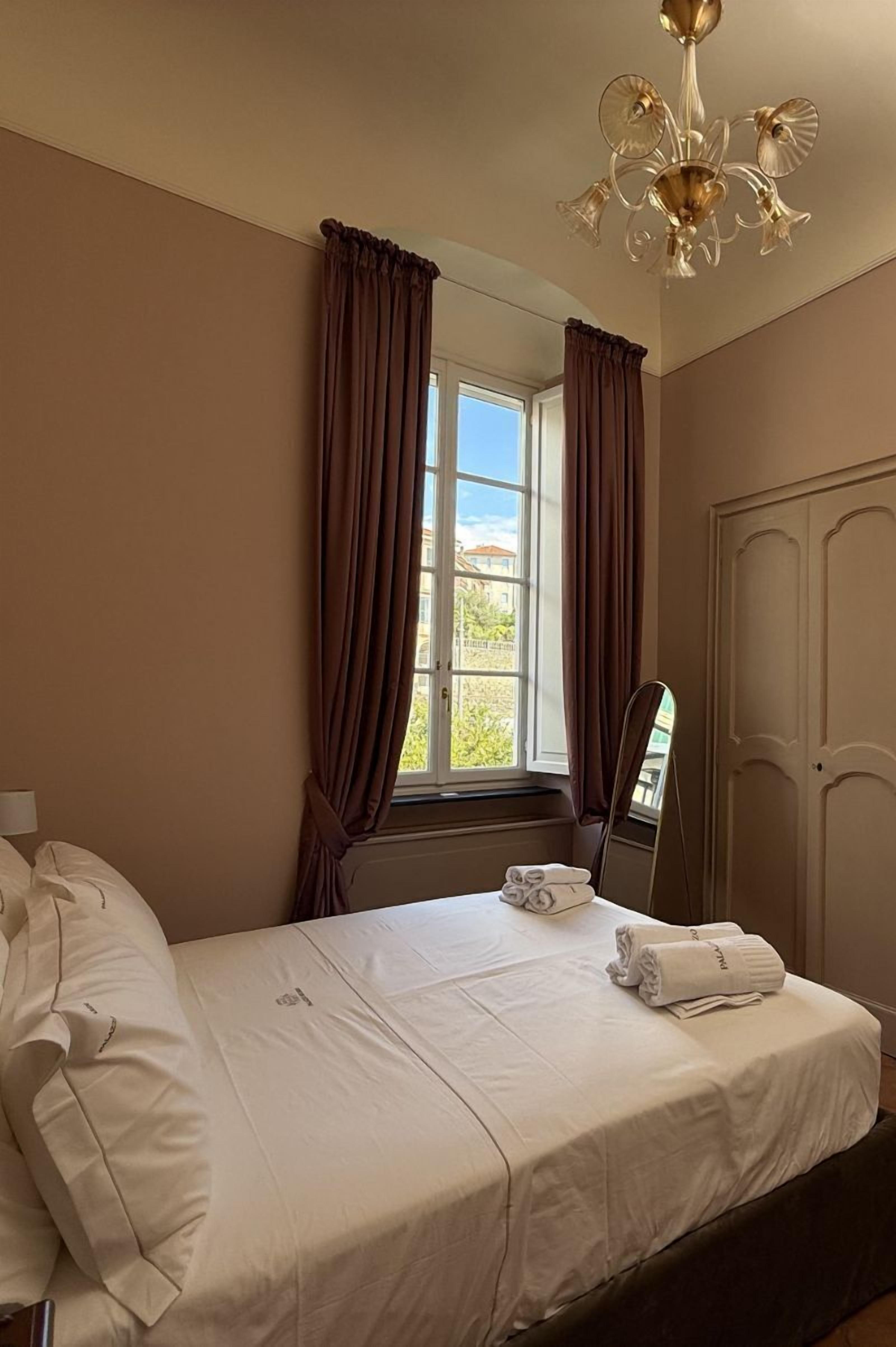 A hotel room with a bed, a window with curtains, a chandelier, and a door.