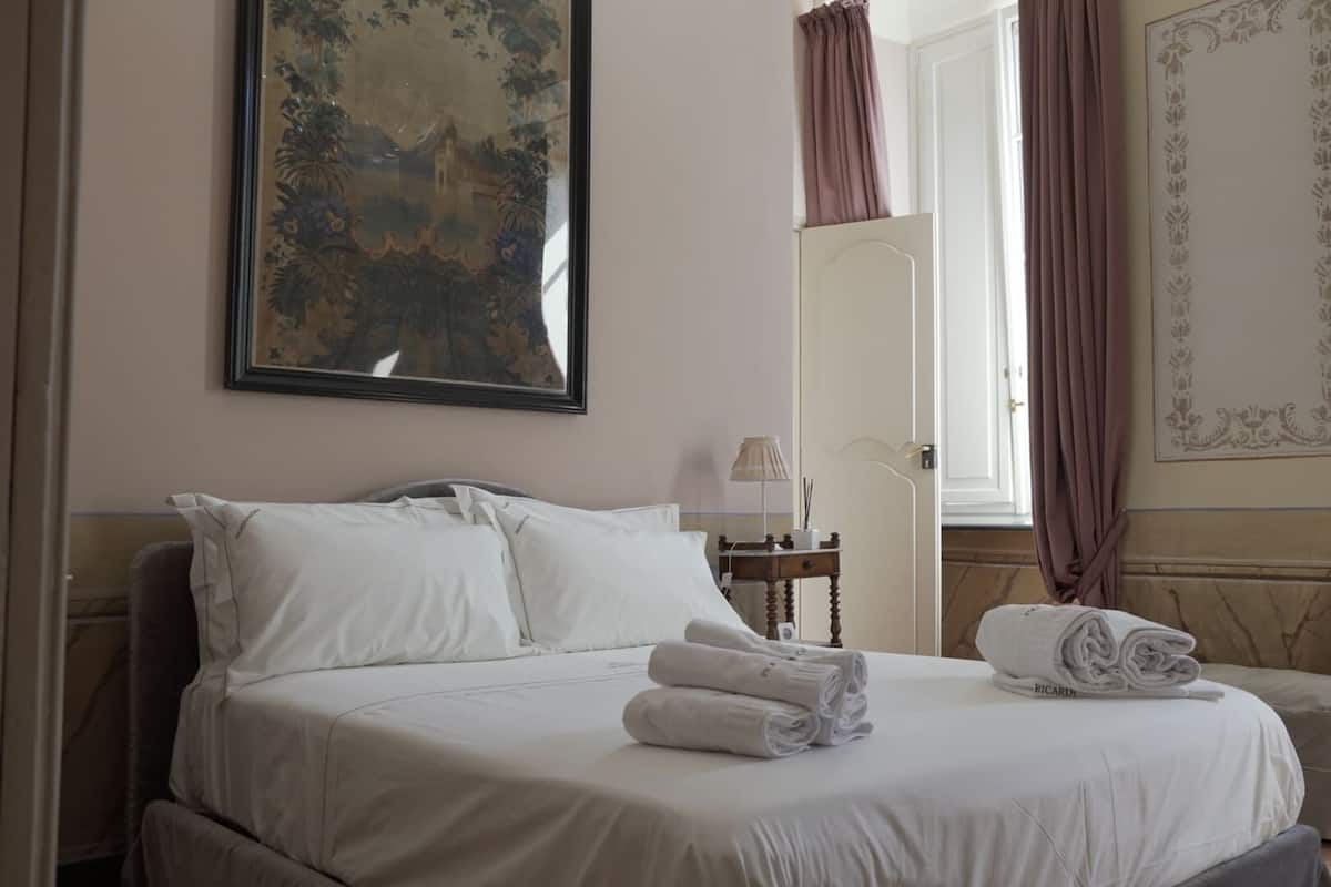 A neatly made bed with white linens and towels, a bedside table with a lamp, and a large framed painting on the wall.