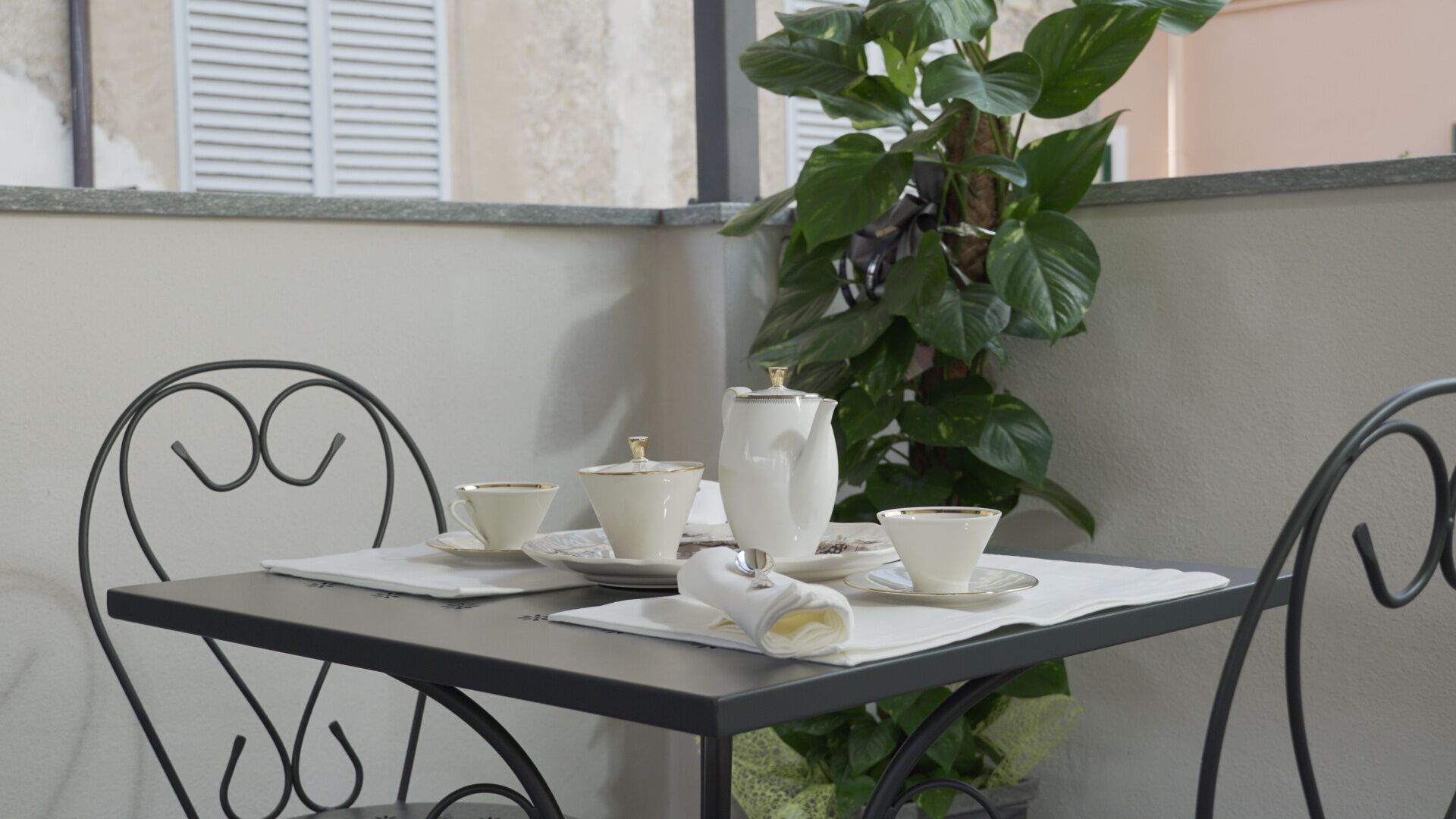 A table set with white porcelain tea service, including a teapot, cups, and saucers, on a balcony with a potted plant and metal chairs.