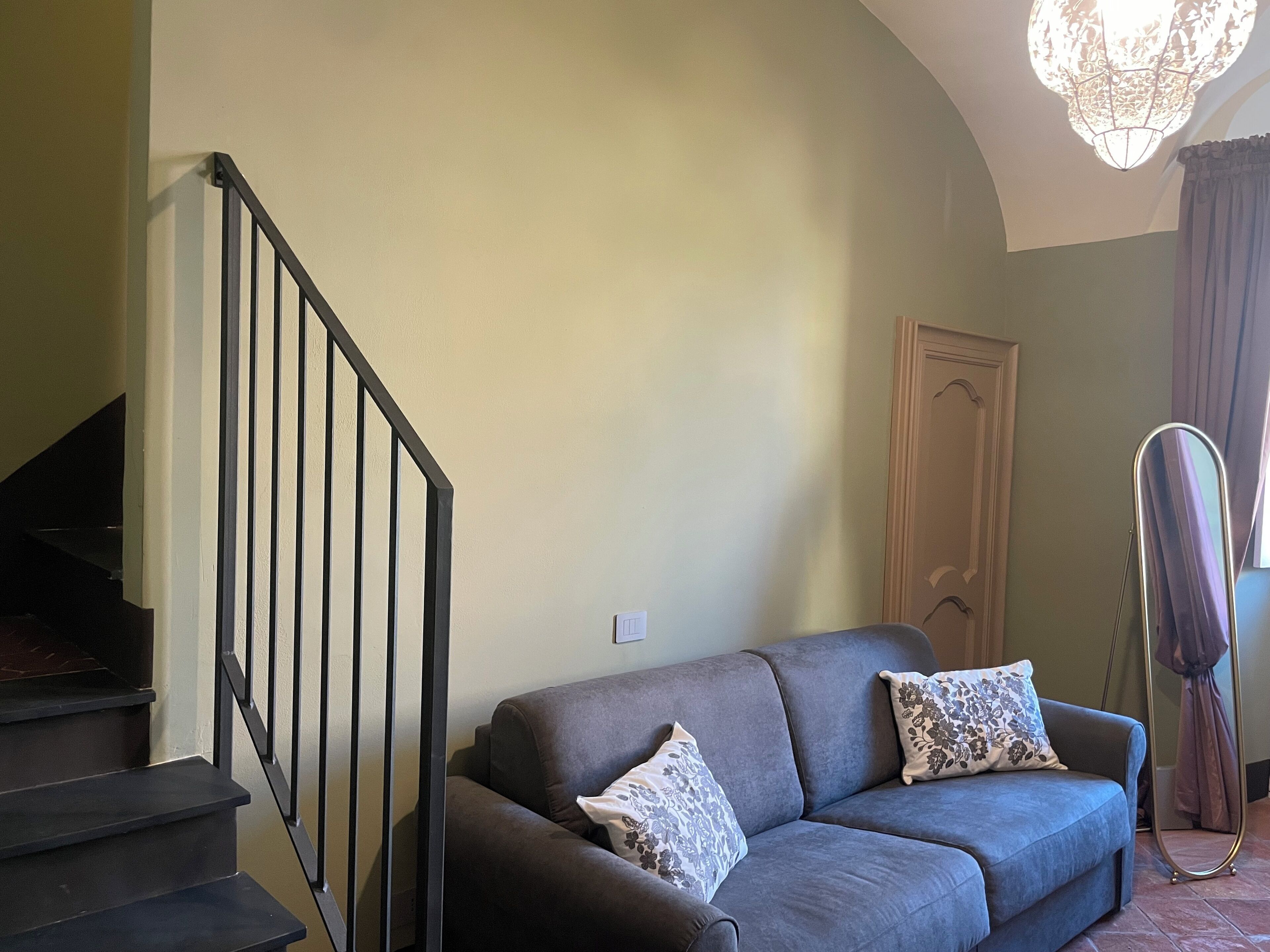 A living room with a grey sofa, two patterned cushions, a black staircase, a wooden door, and a floor lamp.