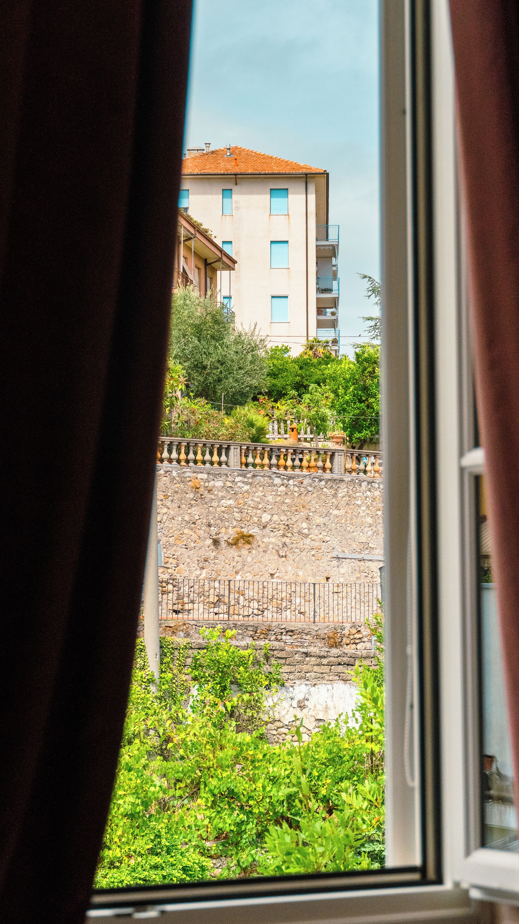 Une vue à travers une fenêtre avec des rideaux, mettant en scène un mur de pierre, de la verdure et un bâtiment doté de nombreuses fenêtres et de balcons.