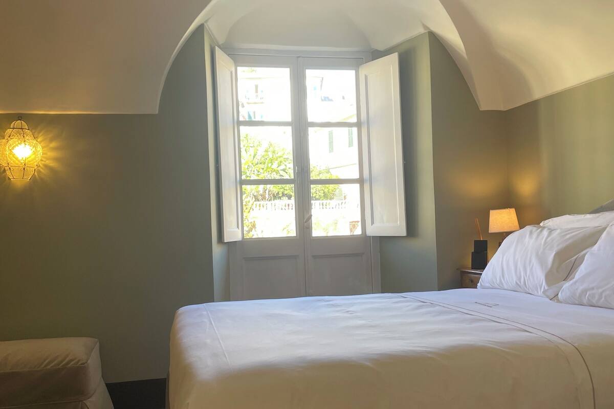 A bedroom with a large bed, a window with open shutters, a wall-mounted lamp, and a nightstand with a lamp.