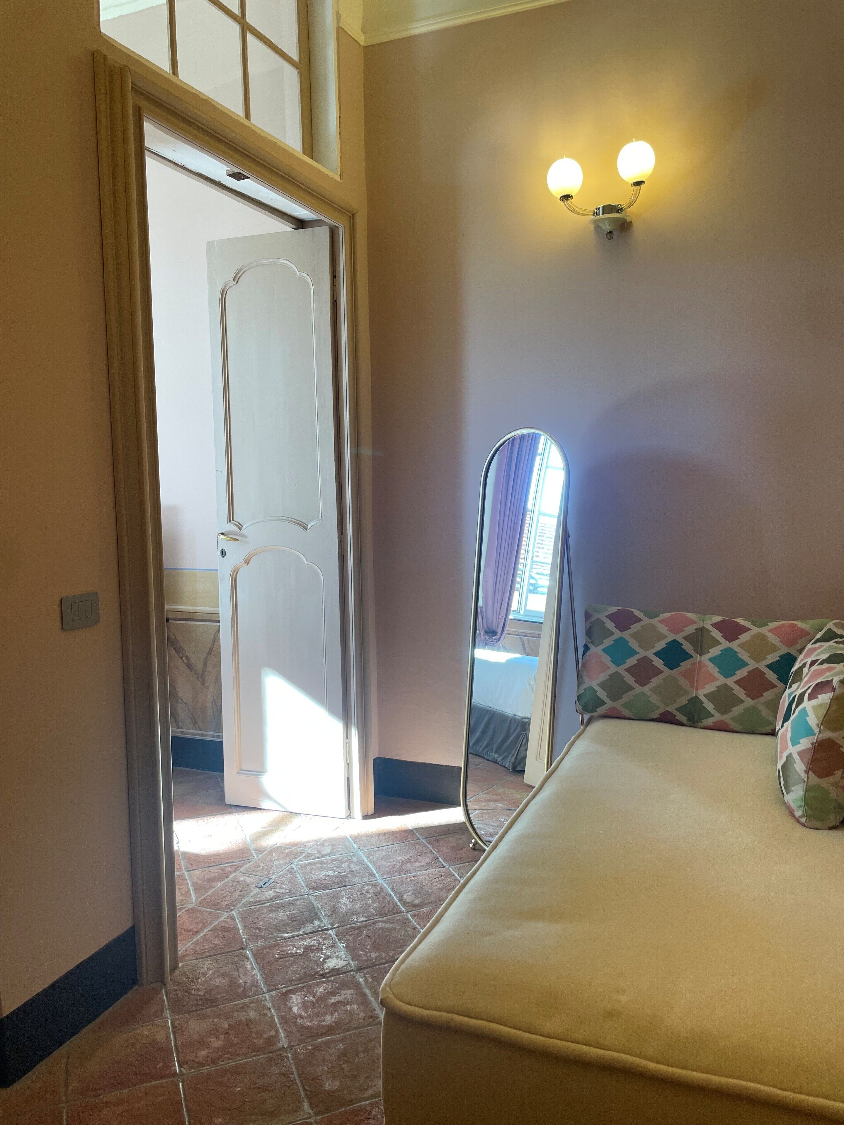 A room with a bed, a mirror, a wall-mounted light fixture, and a door leading to another room.