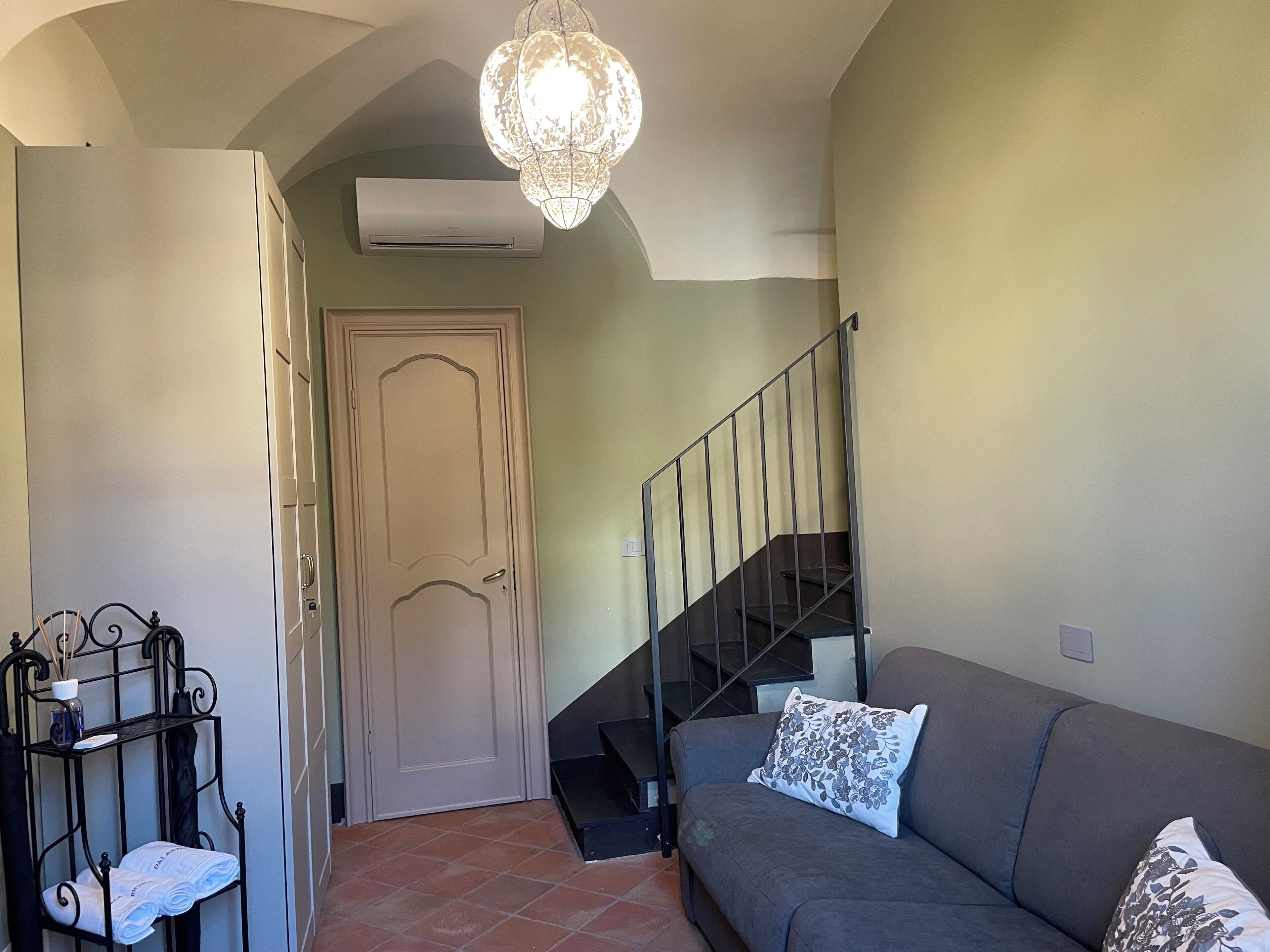 Una sala de estar con un sofá gris, un cojín estampado, una escalera de metal y una puerta con detalles decorativos.