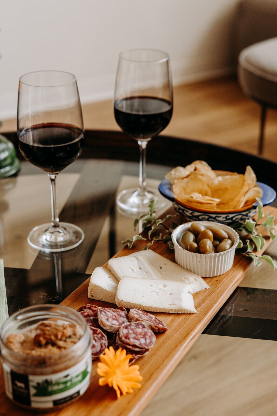 Une planche en bois garnie de fromage, de charcuterie, d’olives et d’un bocal de noix. On peut également apercevoir deux verres de vin rouge.