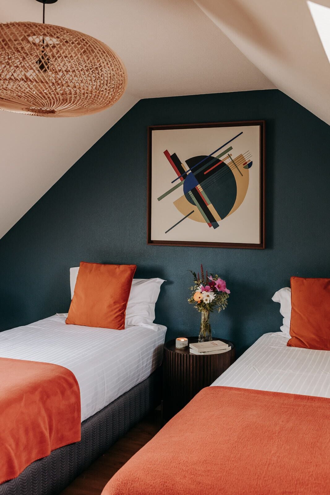 Ein Schlafzimmer mit zwei Betten, einer gewebten Deckenleuchte, einem gerahmten abstrakten Gemälde und einer Vase mit Blumen auf einem Nachttisch.