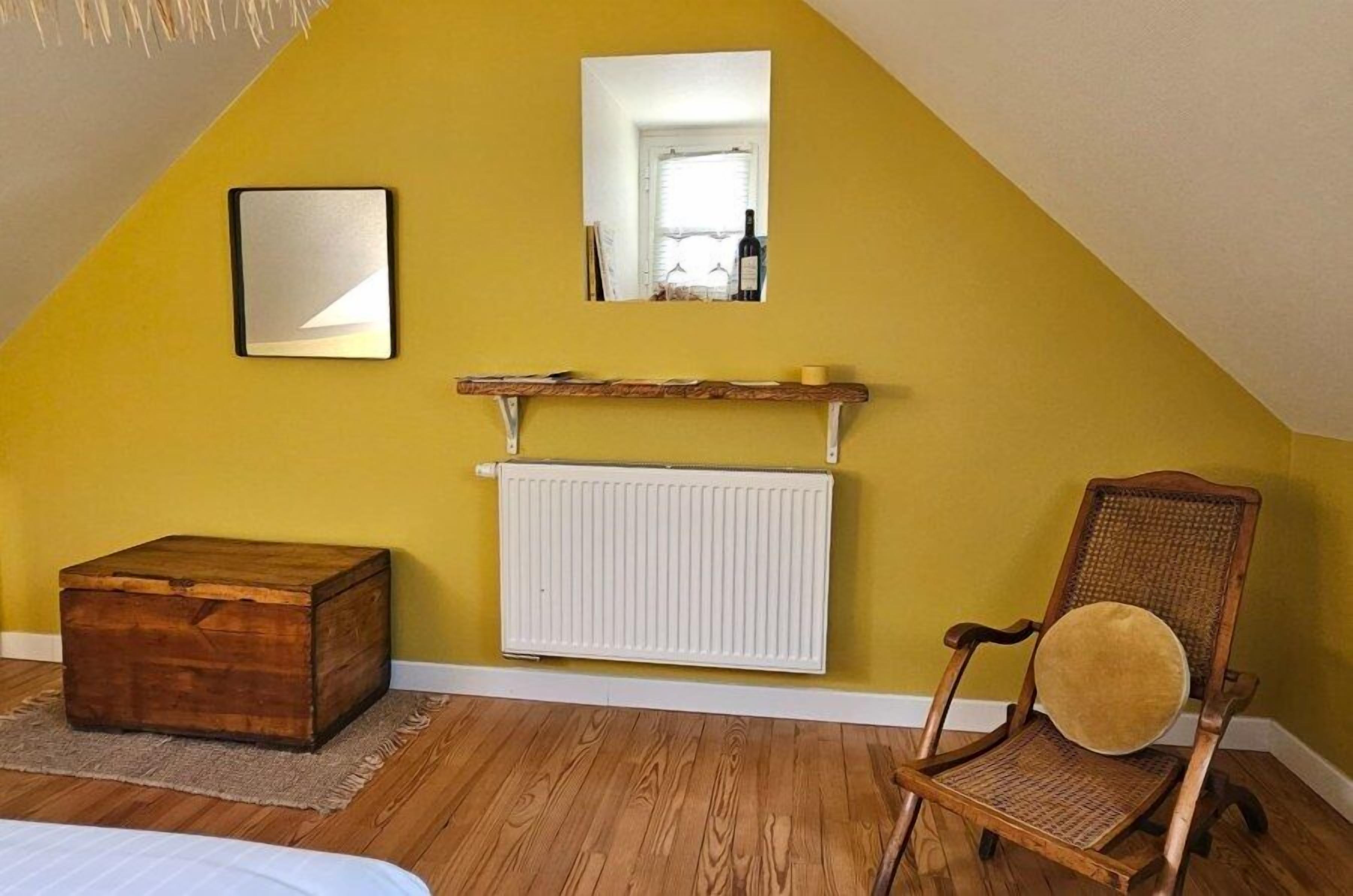 Una habitación con techo inclinado, un aparador de madera, una silla de mimbre, un radiador blanco y un espejo en la pared.
