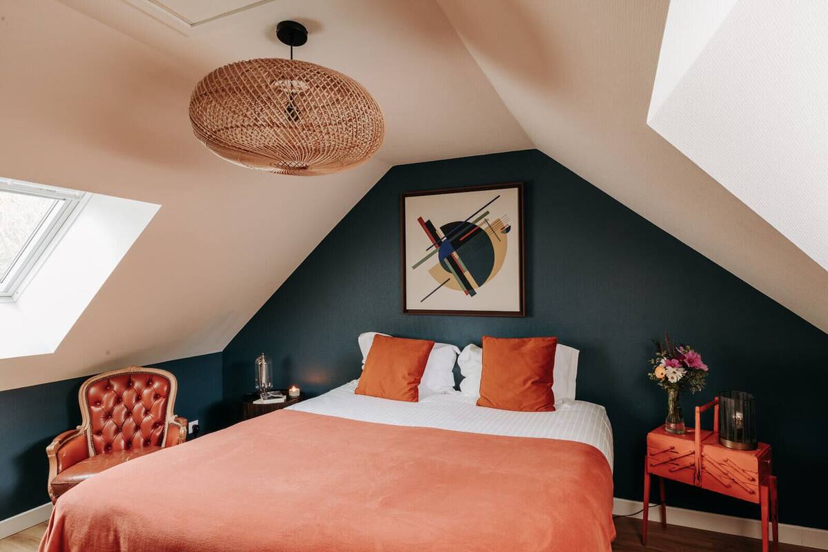 Une chambre à coucher avec un grand lit, un luminaire suspendu, une chaise, une table de chevet et une œuvre d’art accrochée au mur.