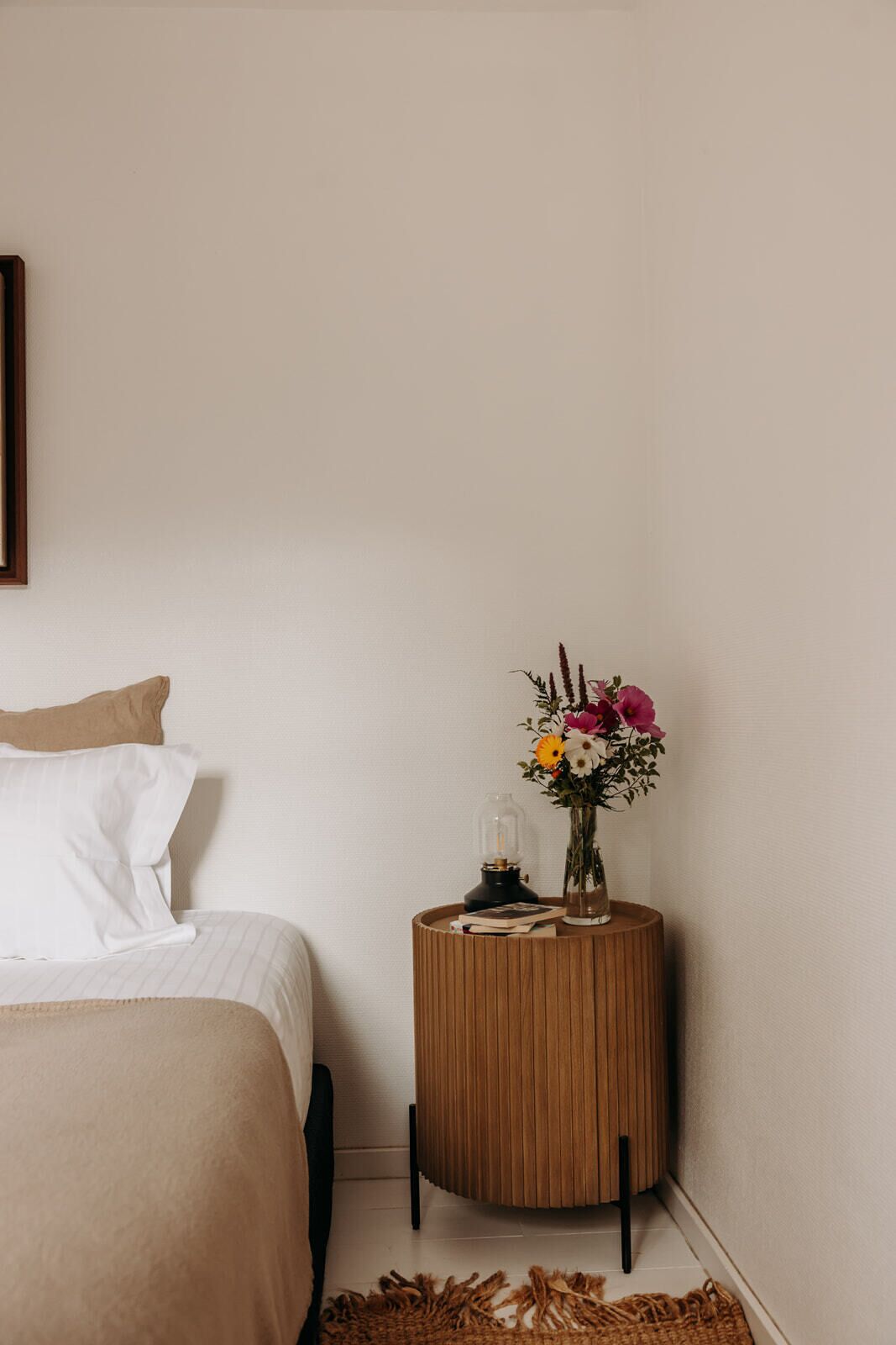 Une chambre à coucher avec un lit, une table de chevet ornée d’un vase de fleurs et une petite lampe.