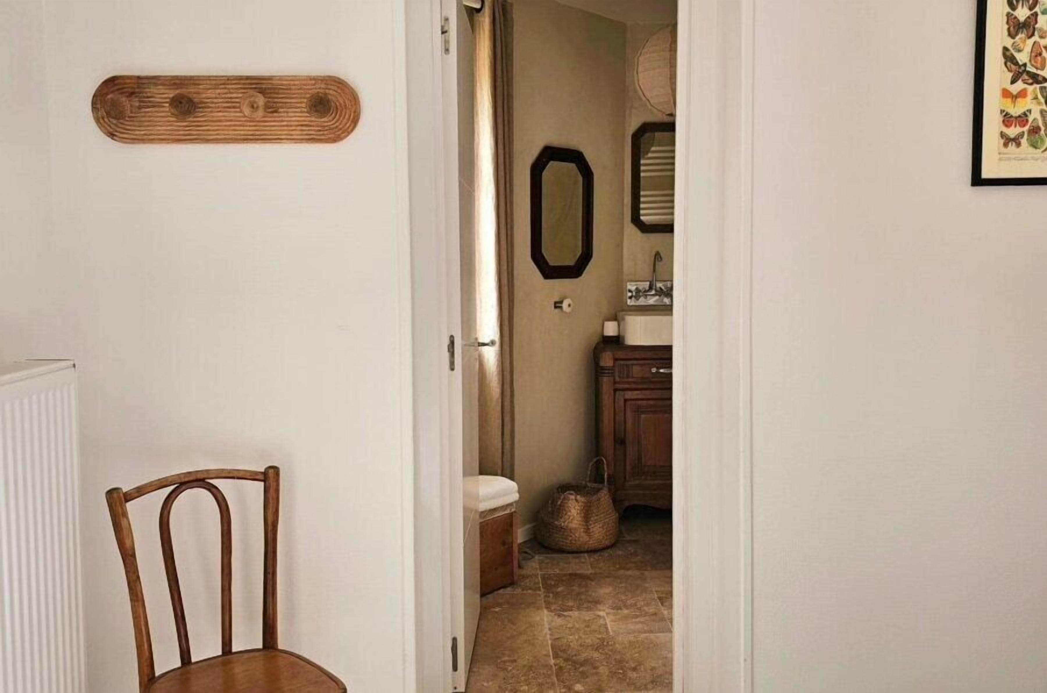 A hallway leading to a bathroom with a wooden chair, a mirror, and a framed butterfly art piece on the wall.