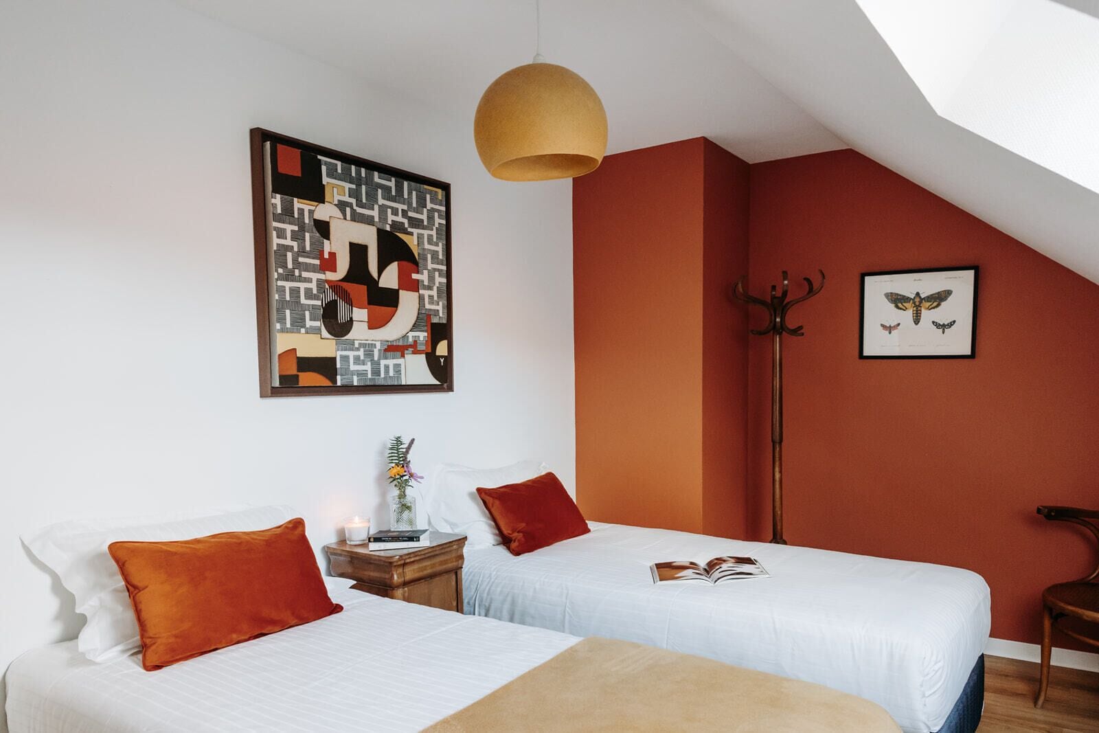 Un dormitorio con dos camas, un lámpara colgante, un cuadro en la pared, un perchero y una imagen enmarcada de una mariposa.