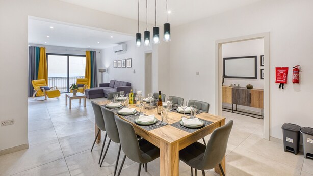 A modern dining area with a wooden table set for six, surrounded by grey chairs. A kitchen area is visible in the background with a microwave and a sink. - Spacious & Cozy 4-Bedroom in Sliema JELL1-2 (Sliema)