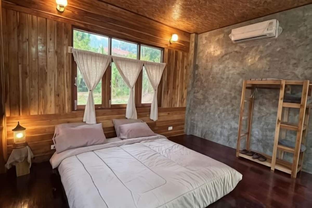 Una habitación de madera con cama, cortinas, una mesita de noche y un estante de madera.