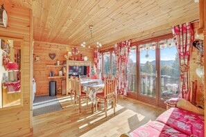 A wooden cabin interior with a dining area, a sofa, and a view through large windows.
