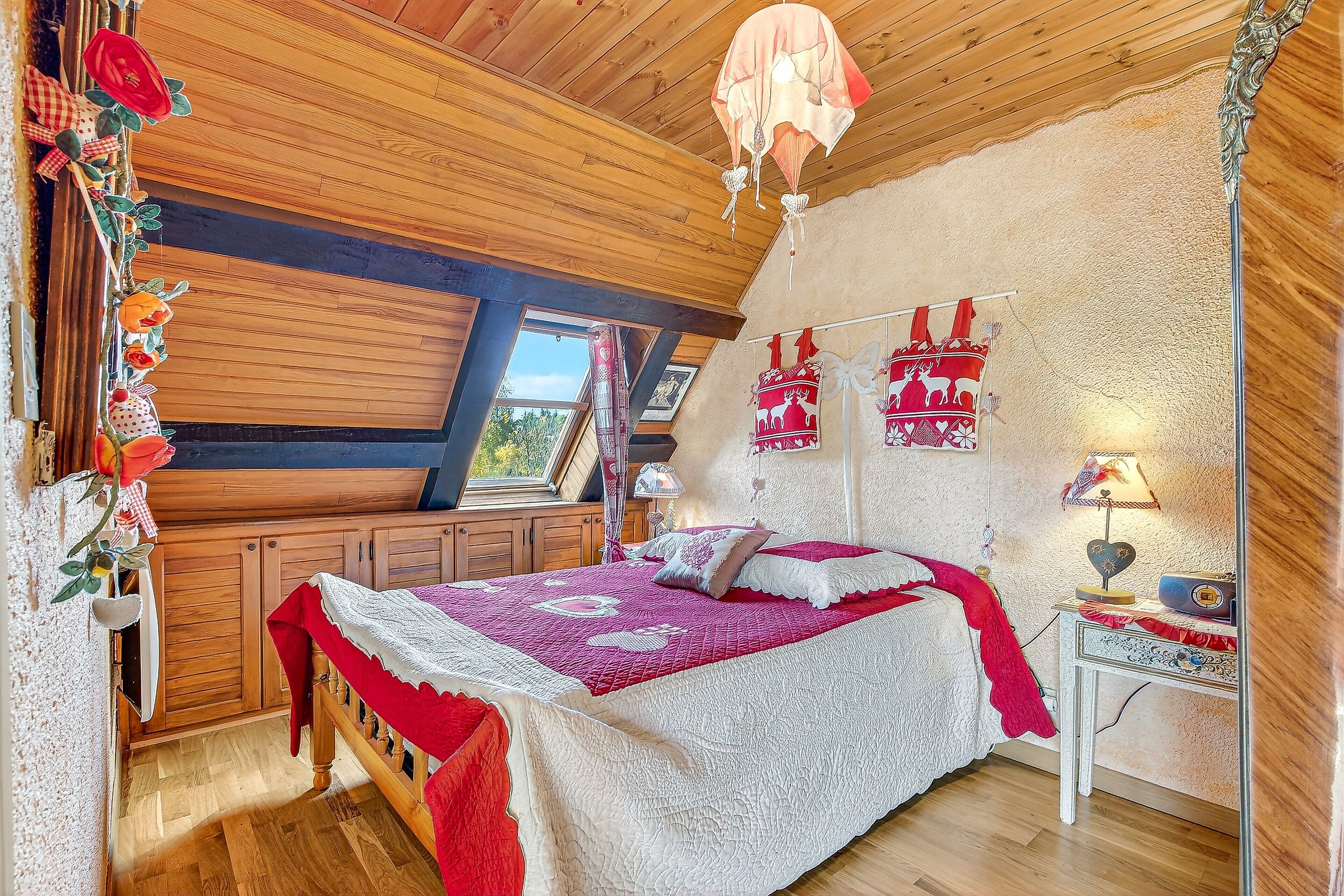 Quarto aconchegante com teto de madeira, uma cama grande com colcha vermelha e branca, e uma pequena mesa de cabeceira com abajur.